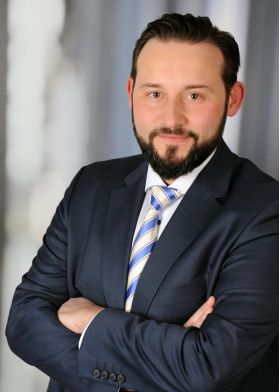 Ein Mann mit dunklen Haaren ist professionell gekleidet und hat die Arme überkreuzt.