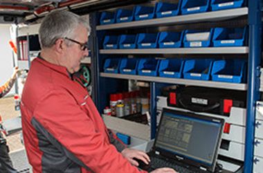 Ein Mann mit roter Jacke steht vor einem Regal und arbeitet an einem Computer.