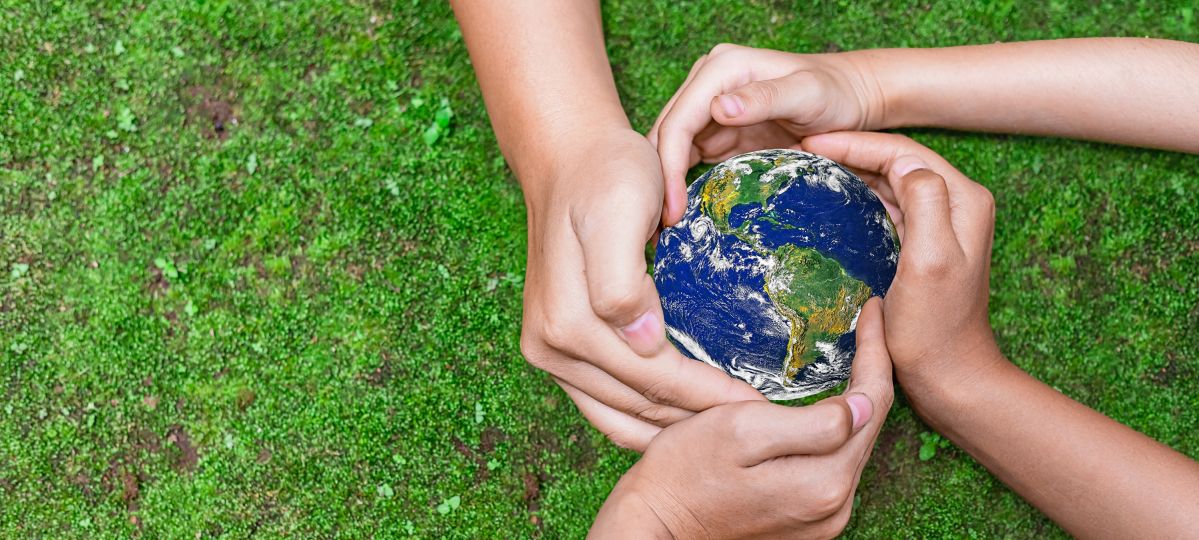 Vier Hände sind um einen kleinen Erdball geschlungen. Im Hintergrund Rasen.