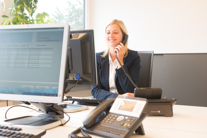 Eine Frau sitzt am Schreibtisch und hat ein Telefon ans Ohr gehalten. Sie ist umgeben von Computer-Bildschirmen.