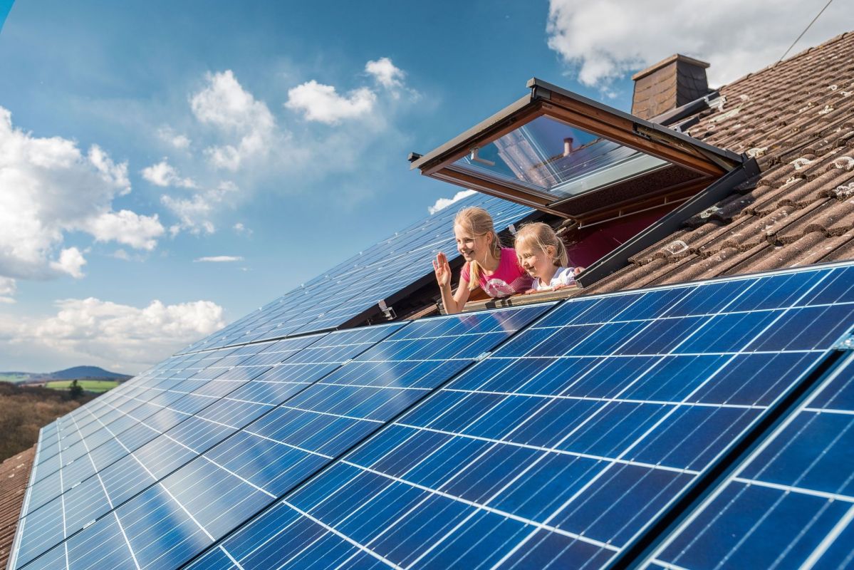 PV-Anlage auf Dach mit 2 Kindern die aus dem Fenster schauen. Im Hintergrund ein sonniger Himmel.