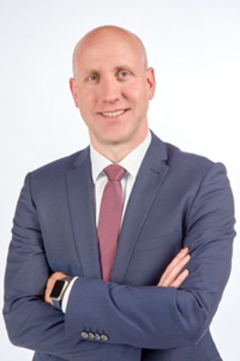 Ein professionell gekleideter Mann hat die Arme verschränkt und lächelt freundlich.