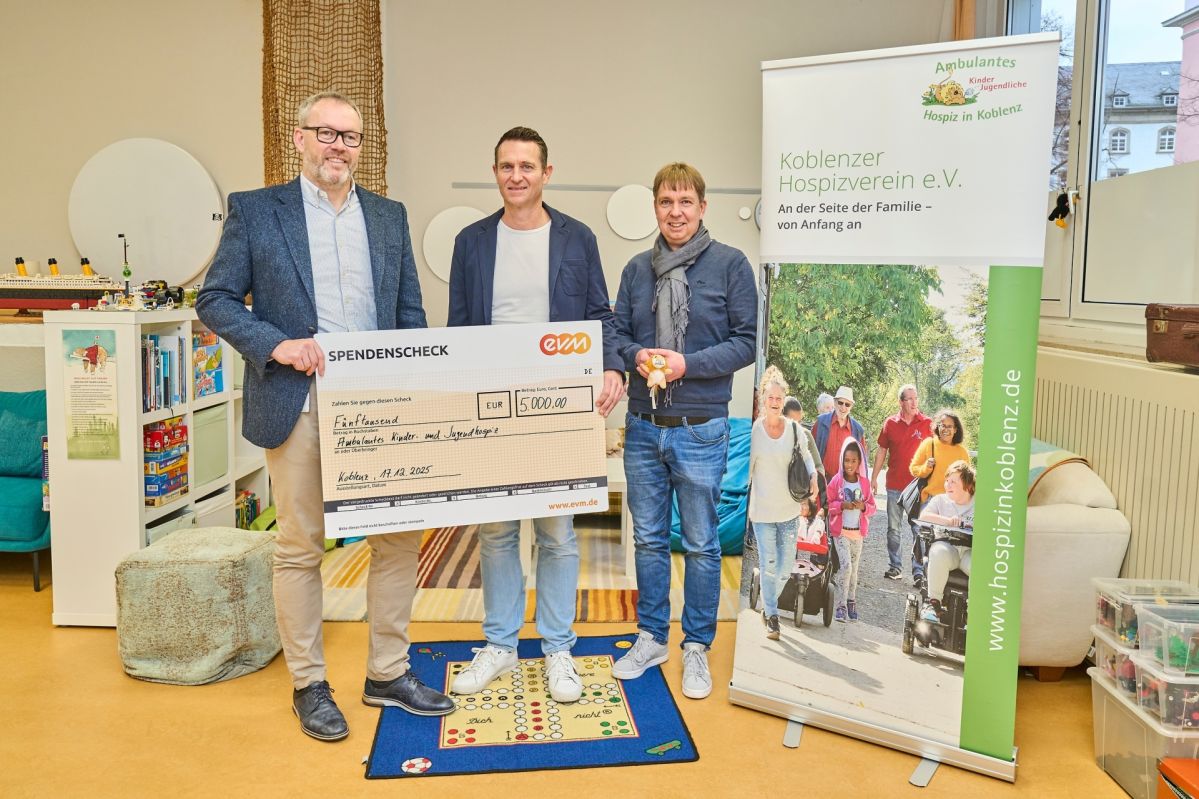 Drei Personen präsentieren in einem hellen Raum einen großen Spendenscheck der evm, neben einem Roll‑up des Koblenzer Hospizvereins e.V.