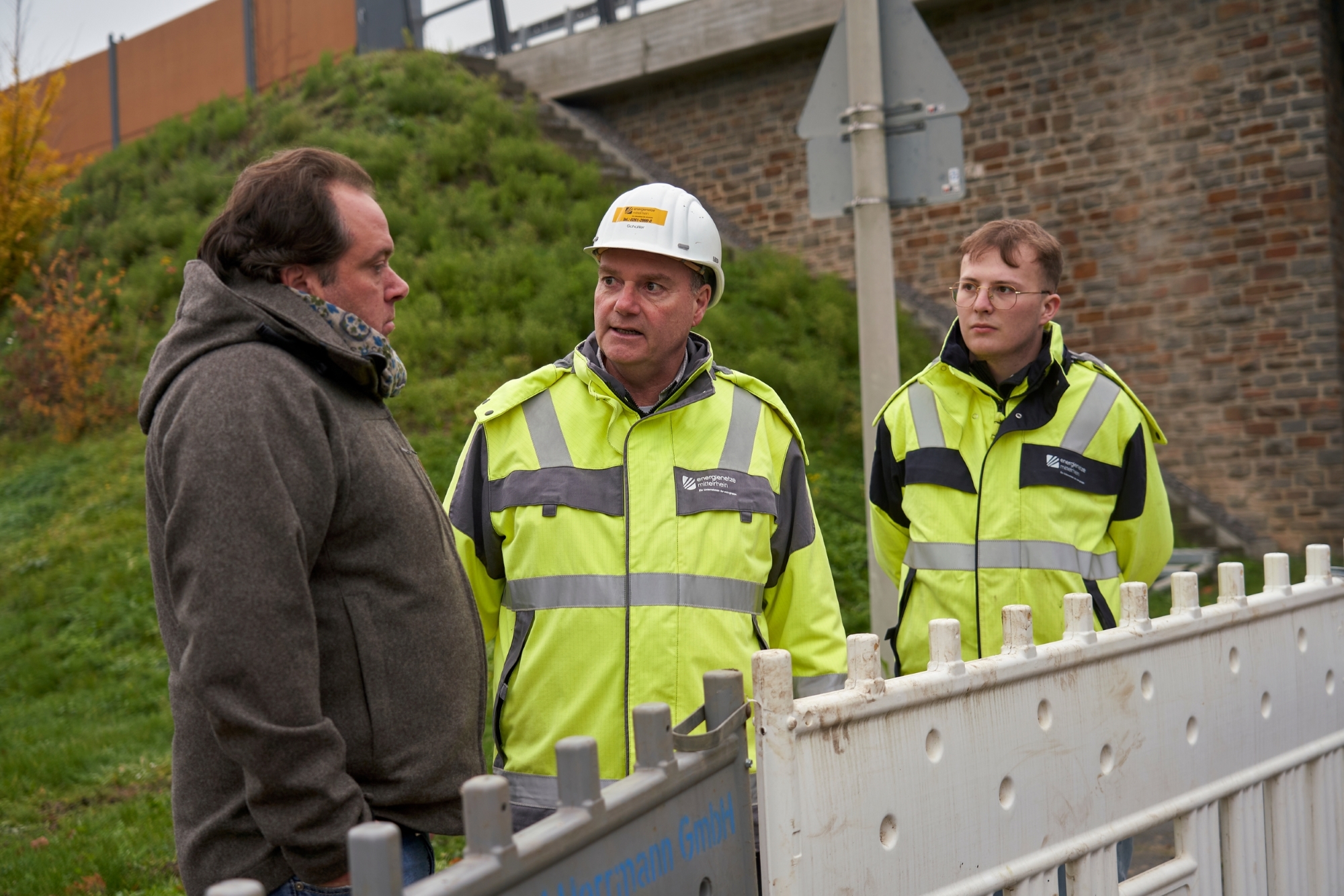 Drei Männer stehen vor einer Absperrung. Zwei von ihnen tragen Schutzkleidung.