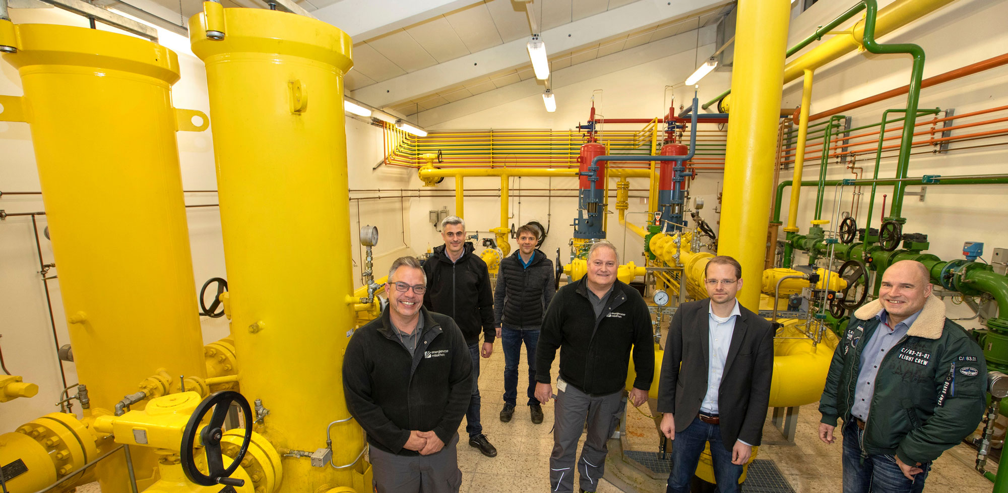 Sechs Männer stehen in einem Raum mit großen gelben Rohren und lächeln freundlich