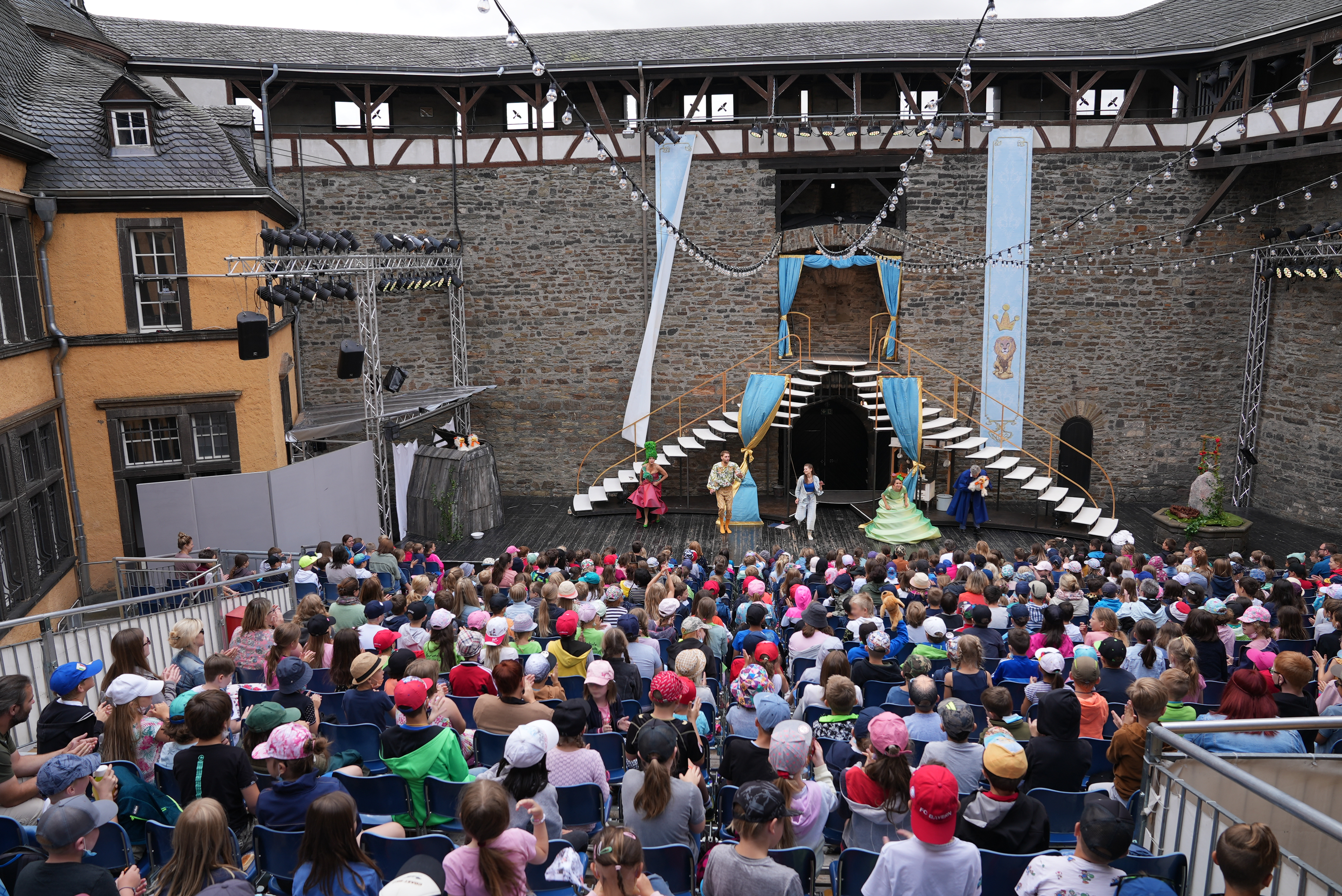 Eine Bühne mit Schauspielern steht vor einer Burgmauer. Im Publikum sitzen viele Kinder