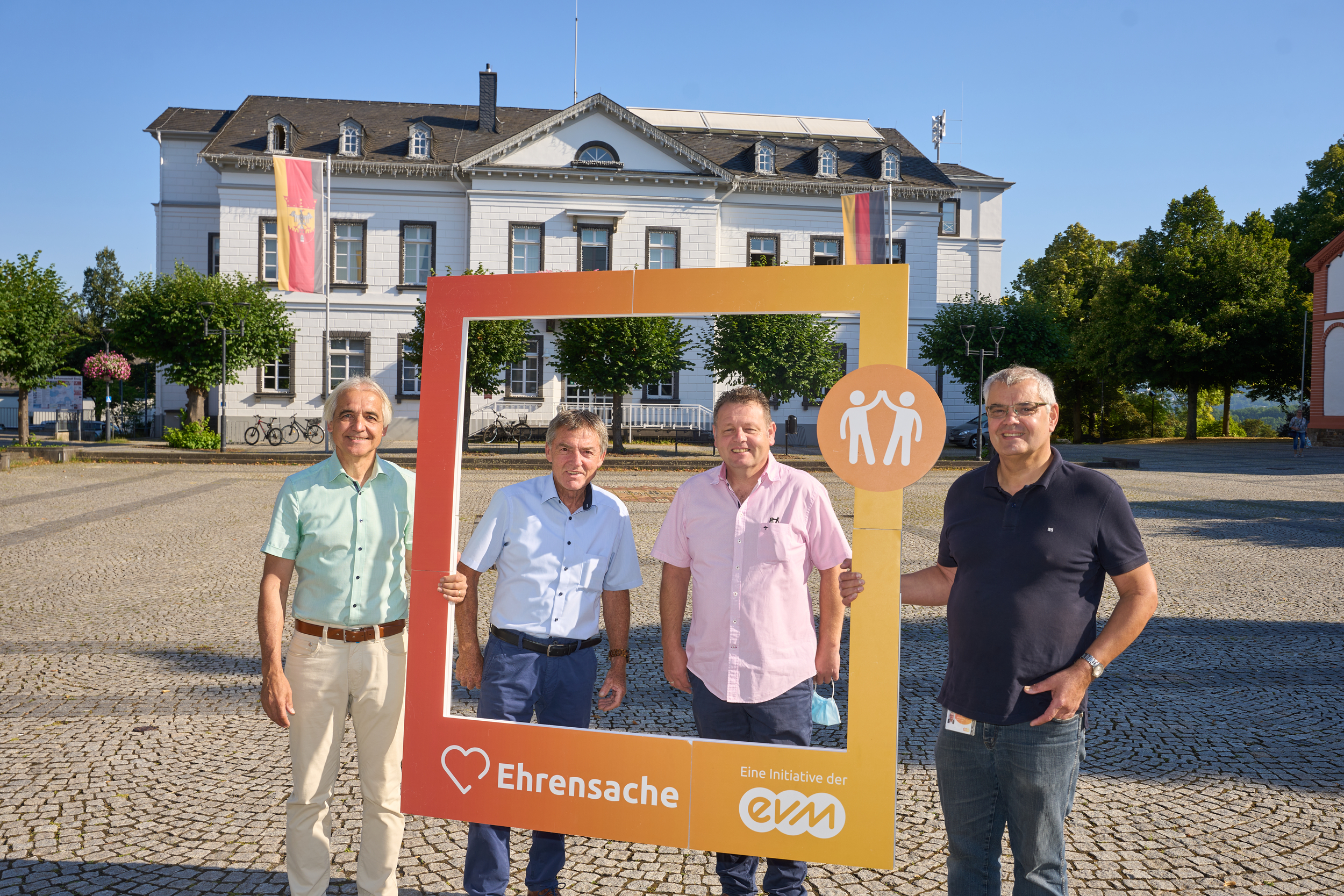 Vier Männer lächeln freundlich und halten einen orangenen Rahmen mit der Aufschrift "Ehrensache".