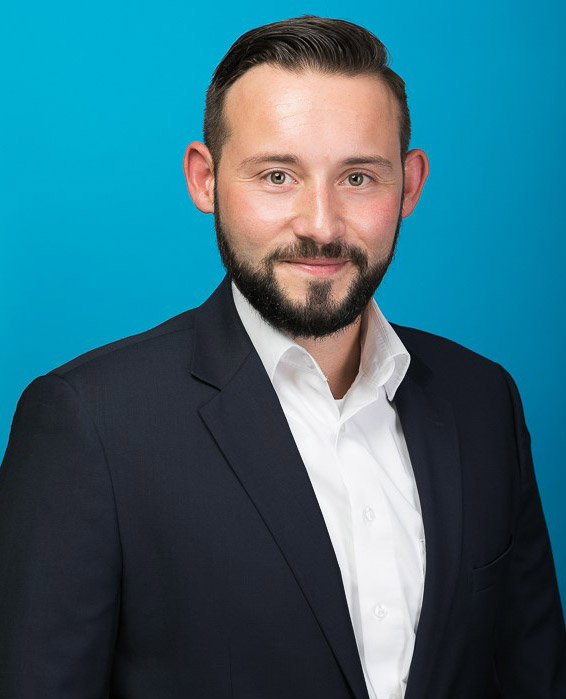 Ein professionell gekleideter Mann steht vor einem blauen Hintergrund und lächelt freundlich