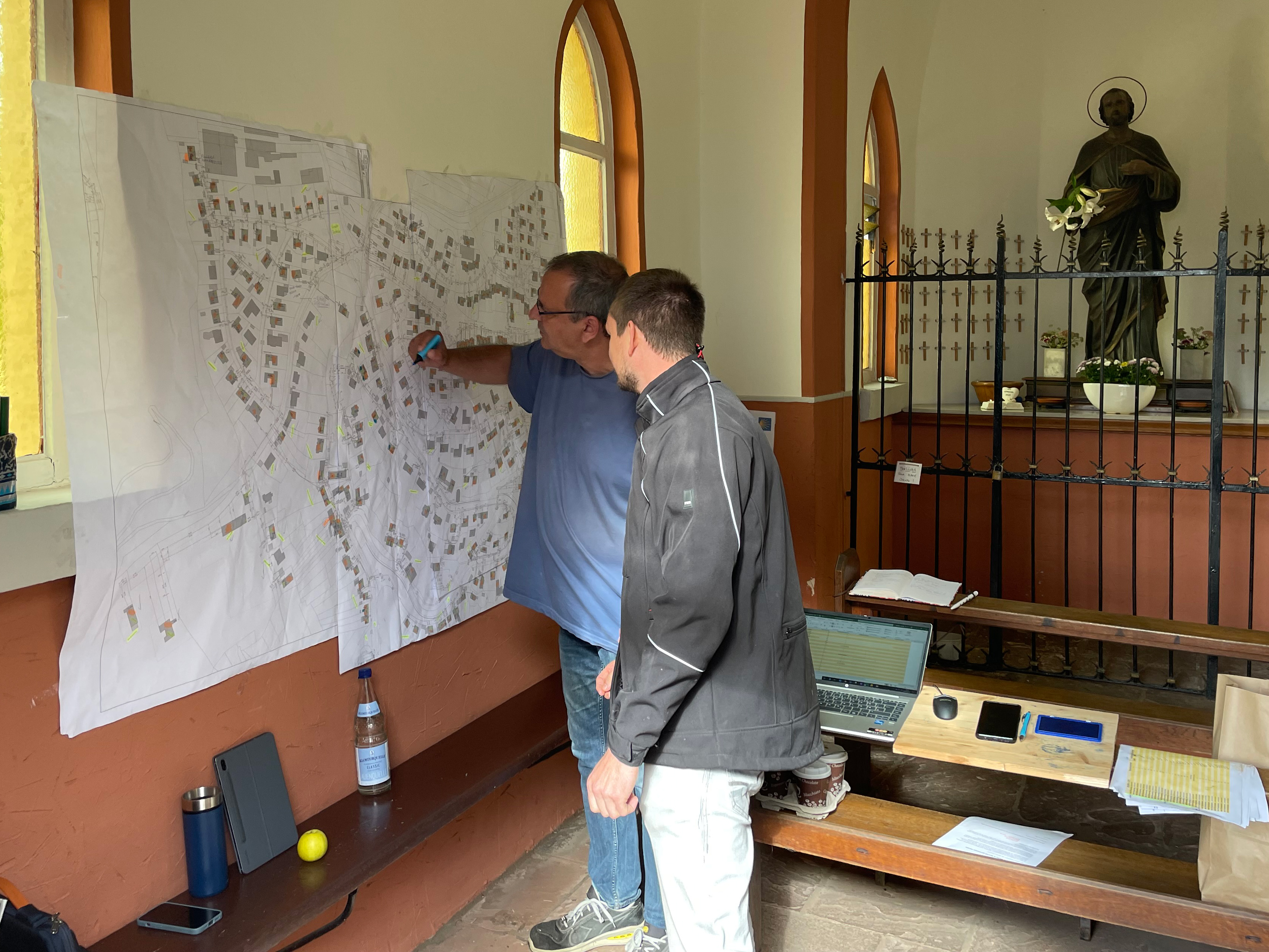 Zwei Männer schauen auf eine Karte die in einer Kapelle aufgehängt ist. 