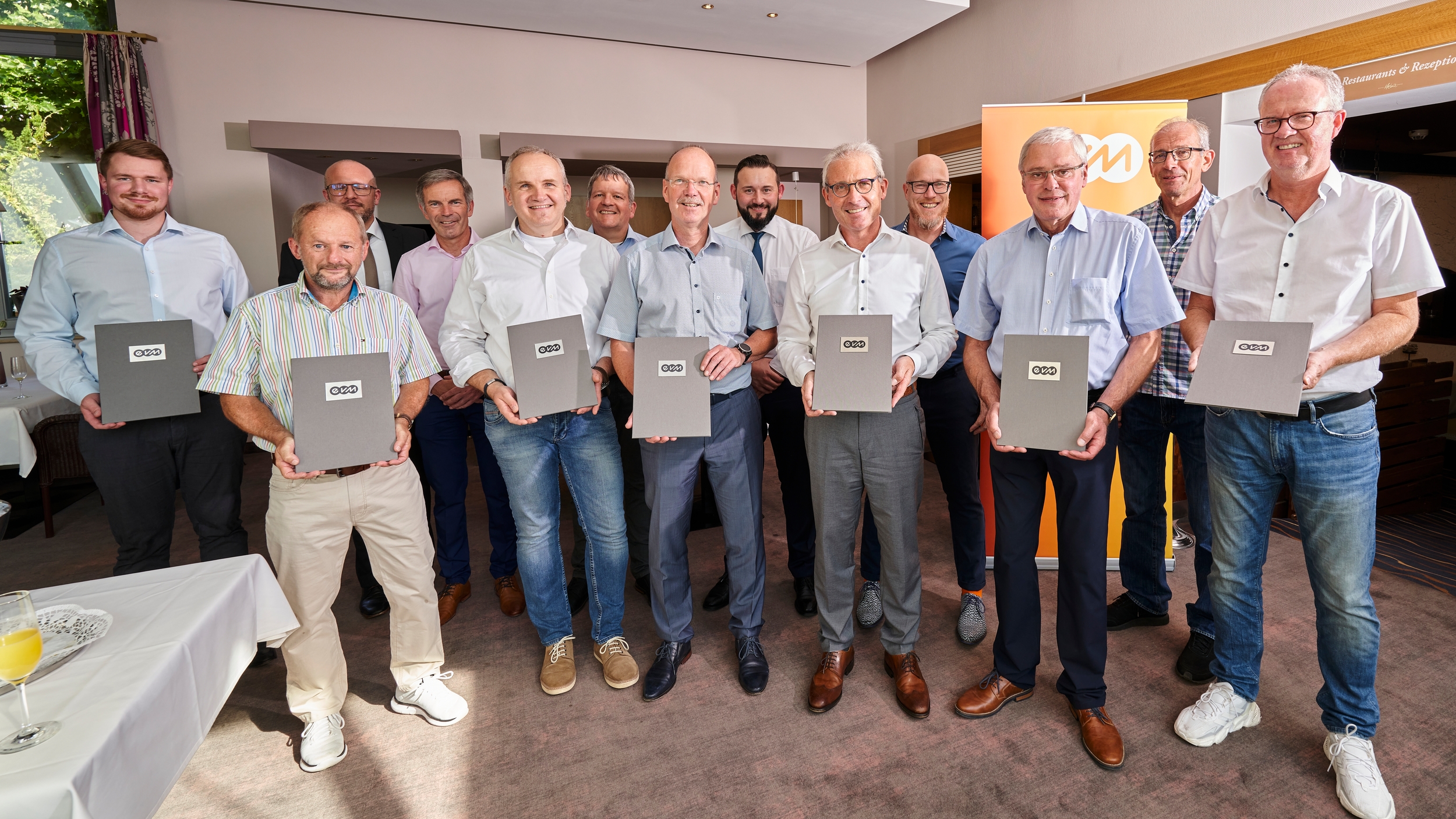 Dreizehn Männer lächeln freundlich und halten graue Mappen mit der Aufschrift "evm".