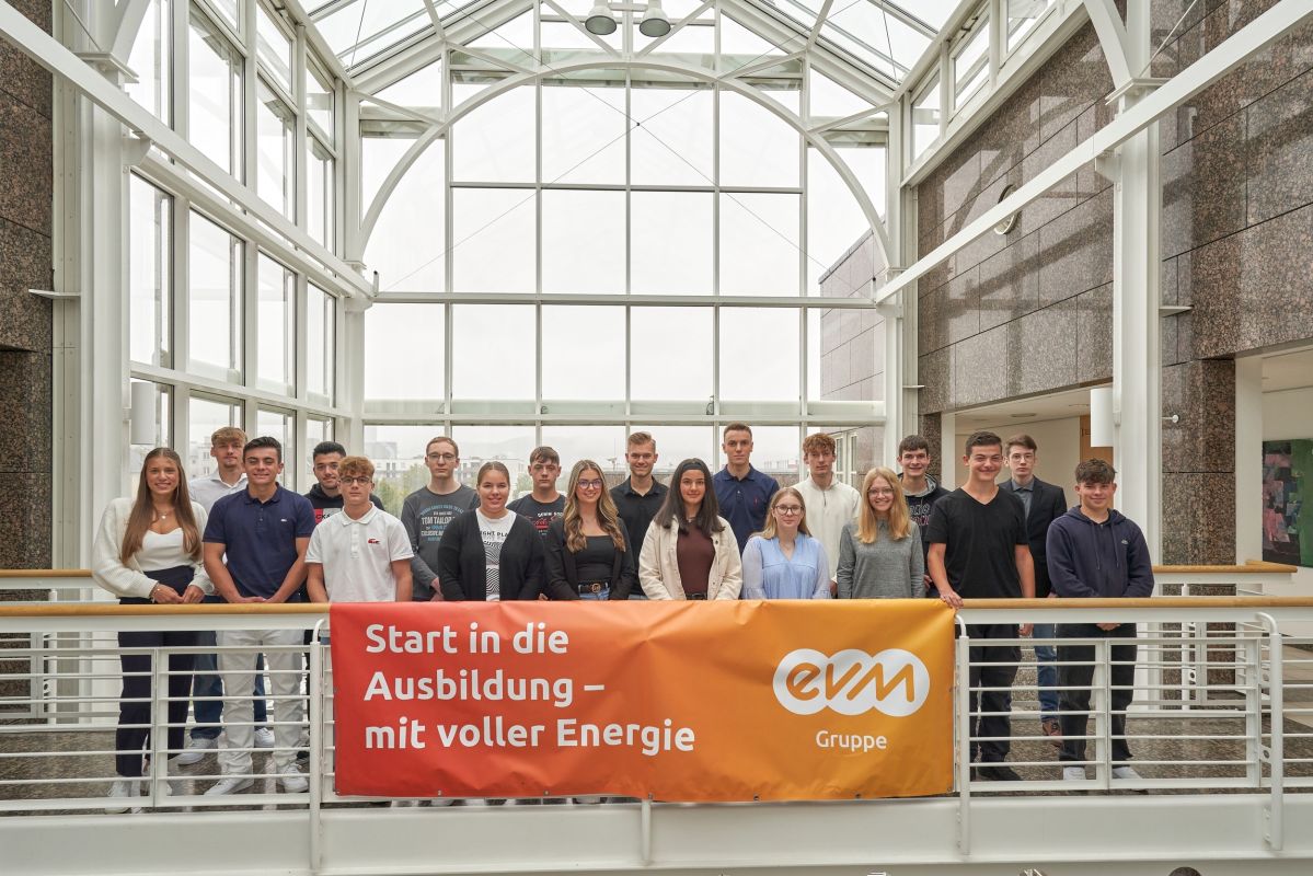 Azubis 2023 in der evm Gruppe Mehrere junge Menschen stehen zusammen auf einem Übergang. Unter ihnen hängt ein orangener Banner mit der Aufschrift "Start in die Ausbildung - mit voller Energie."