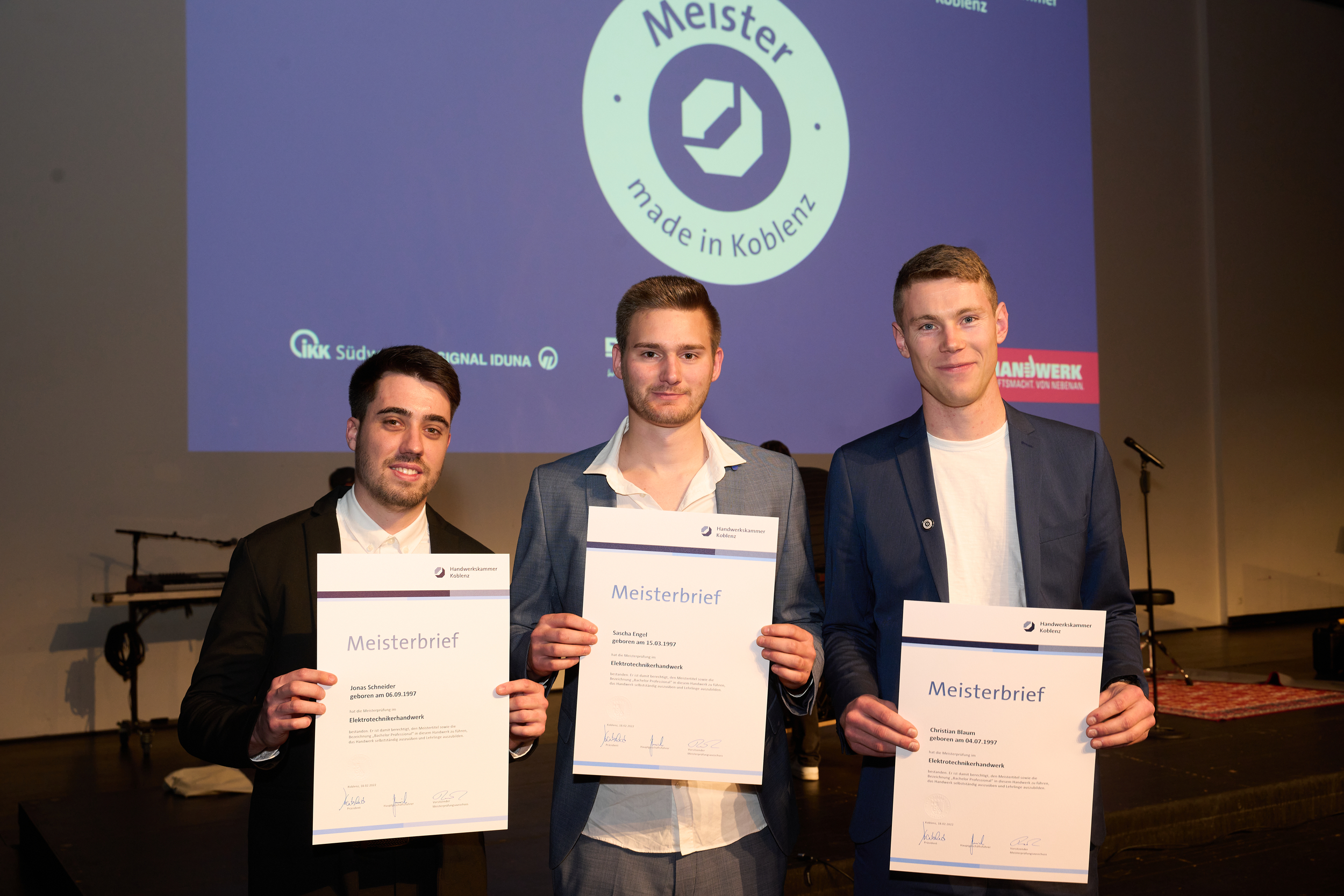 Drei Männer lächeln freundlich und halten Urkunden in der Hand.