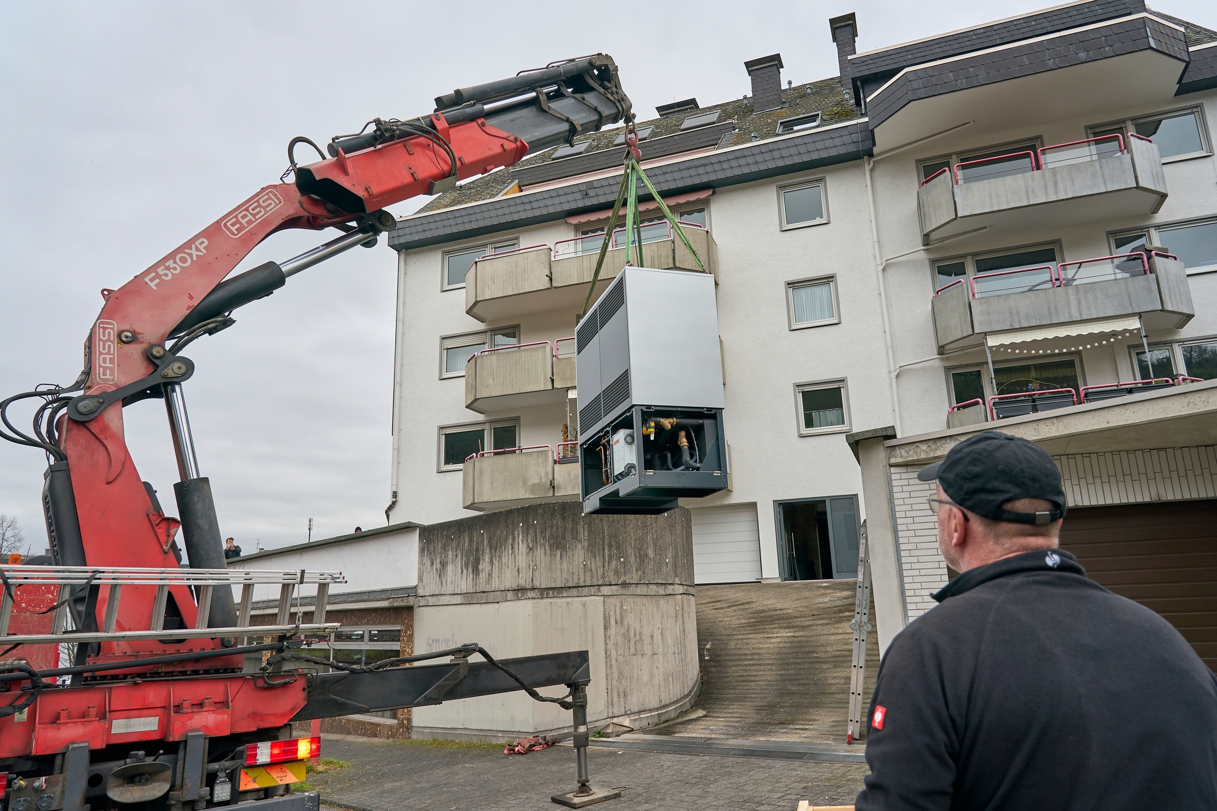 Ein Kran an dem eine Wärmepumpe hängt steht vor einem mehrfamillien Haus