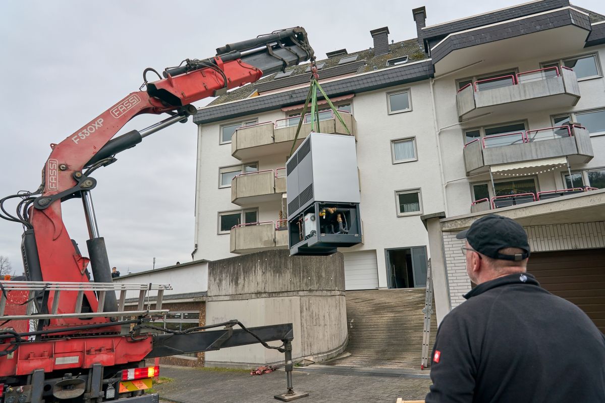 Ein Kran an dem eine Wärmepumpe hängt steht vor einem mehrfamillien Haus
