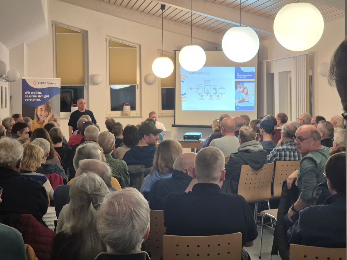 Volle Informationsveranstaltung der KTK in einem hell beleuchteten Raum, in dem zahlreiche Personen auf Stühlen sitzen und einer Präsentation auf einer großen Leinwand folgen. Vorne sprechen Referierende, daneben stehen Banner mit Informationen des Veranstalters.