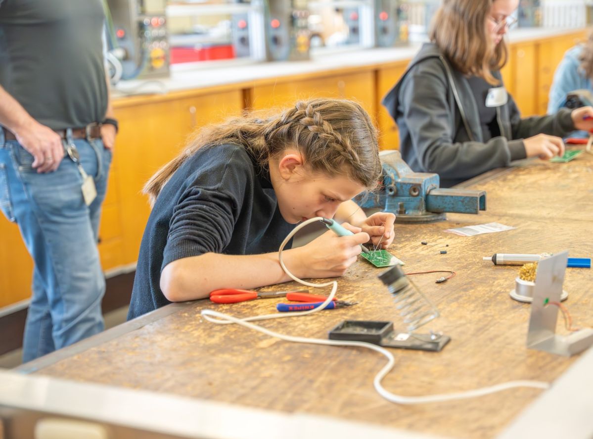 Ein Mädchen mit braunen. geflochtenen Haaren, sitzt in einer Werkstatt und lötet etwas. Um sie herum liegt Werkzeug. Im Hintergrund sind weitere Kinder zu erkennen.