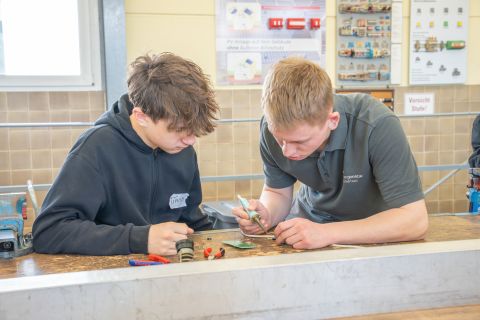 Einem Jungen wird das löten erklärt von einem Azubi. Sie sitzen an einem Tisch in einer Werkstatt. Um sie herum liegt Werkzeug.