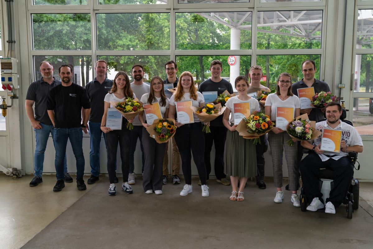 Urkundenübergabe_Azubis-40 Eine Gruppe junger Menschen steht in einer Halle und lächelt freundlich. Sie halten Zertifikate in der Hand und Blumensträuße.