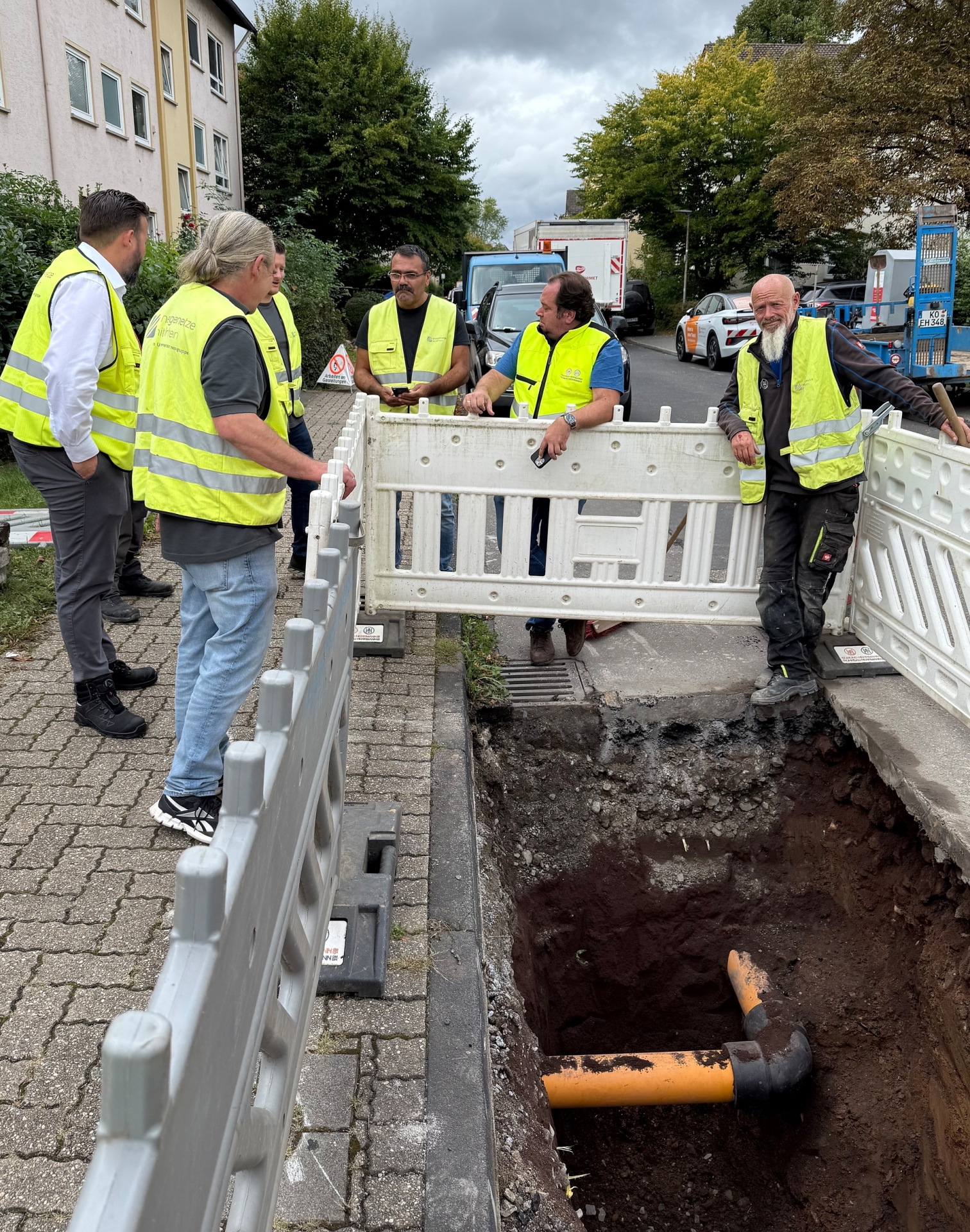 IMG_2526 Mehrere Personen in gelben Schutzwesten stehen an einer Baustelle. Ein orangenes Rohr liegt offen in einem Graben.