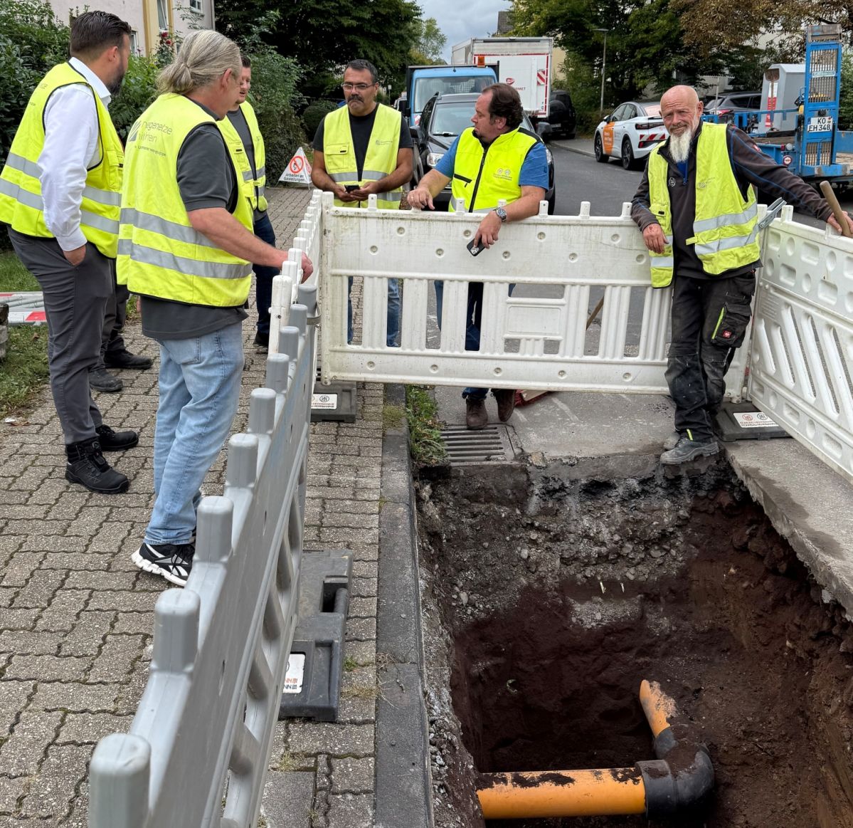 IMG_2526 Mehrere Personen in gelben Schutzwesten stehen an einer Baustelle. Ein orangenes Rohr liegt offen in einem Graben.