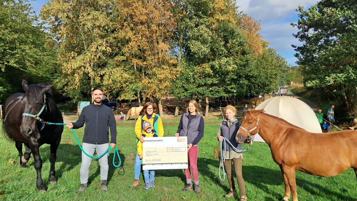evm-Ehrensache Pegasus e.V. Mehrere Personen stehen gemeinsam mit zwei Pferden auf einer Wiese vor einem Zelt und halten einen symbolischen Spendenscheck der evm-Gruppe über 1.000 Euro. Die Spende kommt dem Verein Pegasus e.V. zugute, der sich unter anderem für pferdegestützte Angebote zur Förderung von Kindern, Jugendlichen und Erwachsenen engagiert.