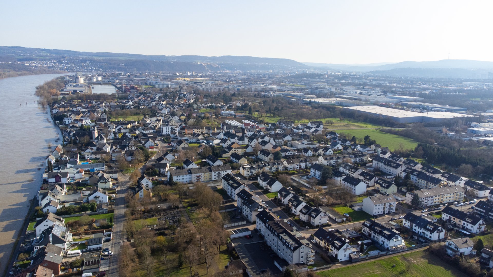 evm-Gruppe schließt Wasserstoff-Pilotprojekt in Koblenz-Kesselheim ab Luftaufnahme von Koblenz-Kesselheim am Rhein mit dicht bebauten Wohngebieten, Grünflächen und Gewerbearealen. Links verläuft der breite Fluss, während sich im Hintergrund Hügel und ein weitläufiges Stadtgebiet erstrecken.