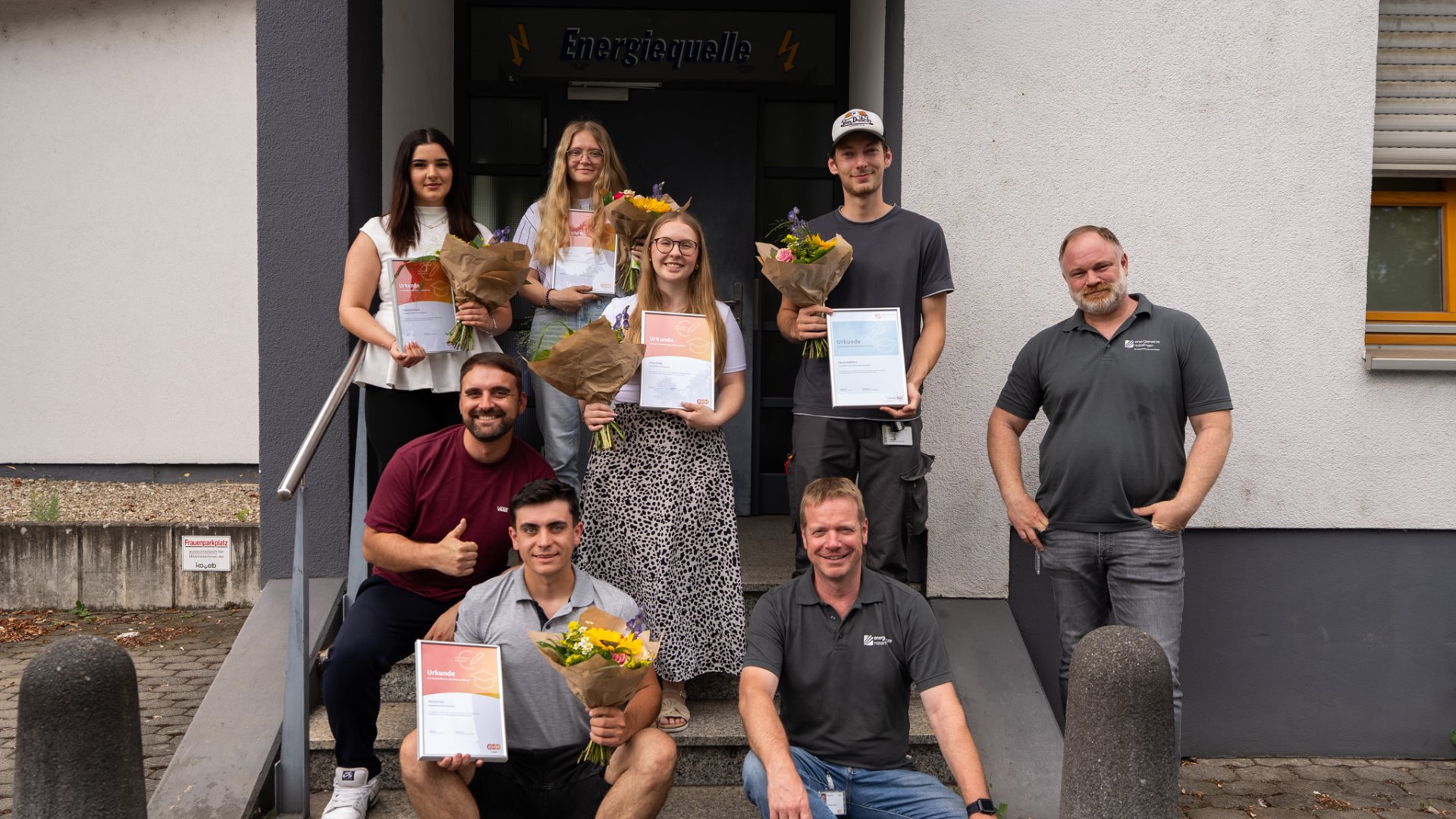 Mehrere Absolventinnen und Absolventen der evm‑Gruppe stehen vor dem Gebäude der Ausbildungswerkstatt „Energiequelle“ und halten ihre Abschlussurkunden sowie Blumensträuße in den Händen. Gemeinsam mit den Ausbildern Nico Sartorius, Joachim Mallmann und Florian Arzbächer freuen sie sich über den erfolgreichen Ausbildungsabschluss.