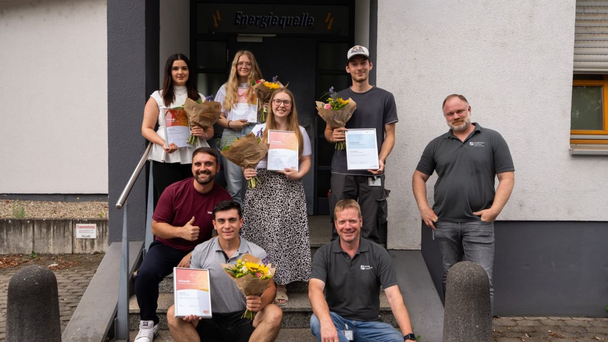 evm-Gruppe übernimmt alle ausgelernten Auszubildenden Mehrere Absolventinnen und Absolventen der evm‑Gruppe stehen vor dem Gebäude der Ausbildungswerkstatt „Energiequelle“ und halten ihre Abschlussurkunden sowie Blumensträuße in den Händen. Gemeinsam mit den Ausbildern Nico Sartorius, Joachim Mallmann und Florian Arzbächer freuen sie sich über den erfolgreichen Ausbildungsabschluss.